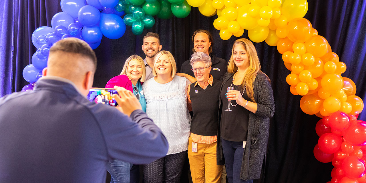 A group of SilverPeak Strategic Capital associates pose for a photo under an arch of rainbow colored balloons at a corporate event
